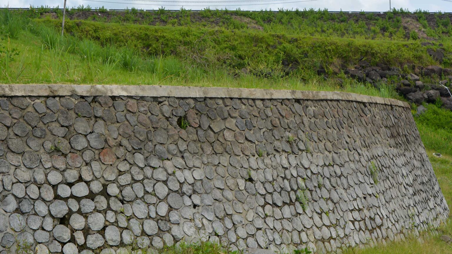 An inclined concrete retaining wall using split face round basalt stones as a facing material.