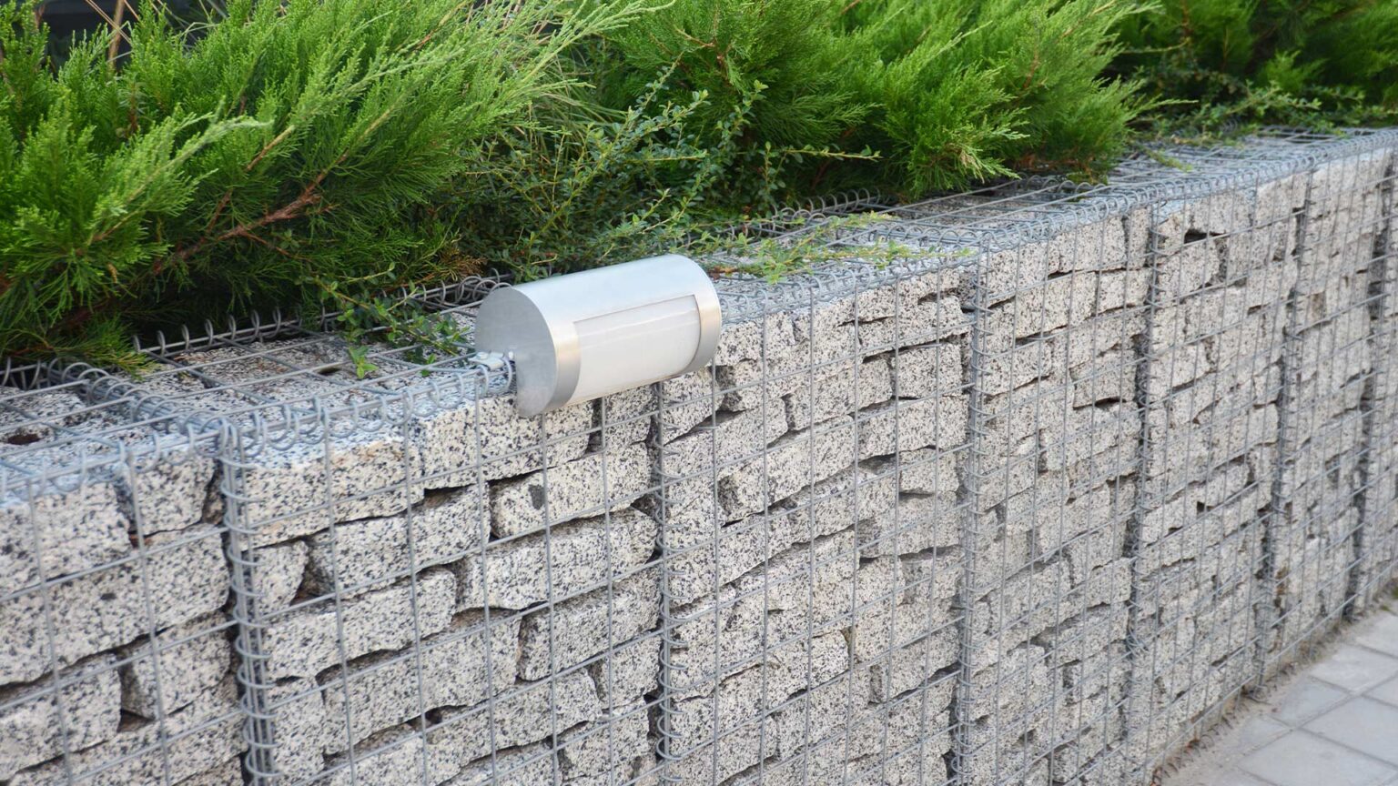 A waist high gabion cage dwarf retaining wall filled with silver granite blocks and fitted with exterior lights.